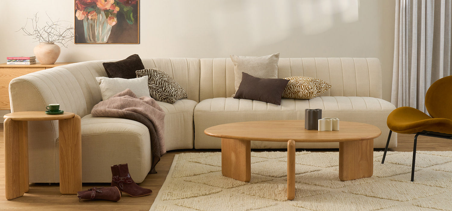 Modern living room with a beige sofa and green cushions beside a wooden slatted console table, decorated with a black vase, white sphere, and oval mirror above.