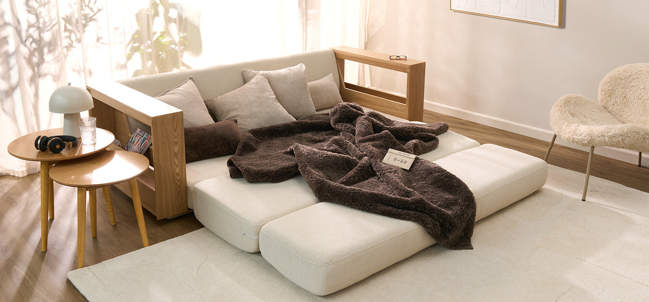 Modern sofa bed styled with neutral cushions and throw blanket, set in a cozy bedroom with woven rug, shelf, and decorative accents.