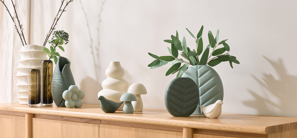 Collection of modern ceramic vases in black, white, terracotta, and textured finishes displayed on a wooden table with dried floral stems.