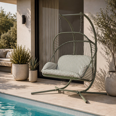 A green metal hanging egg chair with striped cushions sits on a patio beside a swimming pool. The chair is styled in an outdoor residential setting with potted plants and a modern building facade in the background.