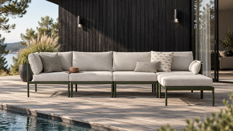 A modern outdoor sectional sofa with light beige cushions and dark green metal frames, arranged on a wooden deck beside a swimming pool in front of a dark wood-paneled house.