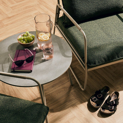 A styled interior scene with dark green upholstered chairs, a round gray side table holding a glass pitcher of lemonade, sunglasses, a bowl of green grapes, and a purple notebook, plus a pair of black slide sandals on a light wood herringbone floor.
