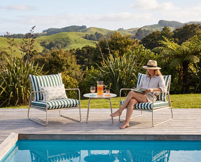 A woman sits barefoot on a striped outdoor lounge chair beside a pool, reading a book on a small round table with two matching chairs and a garden backdrop of rolling green hills.
